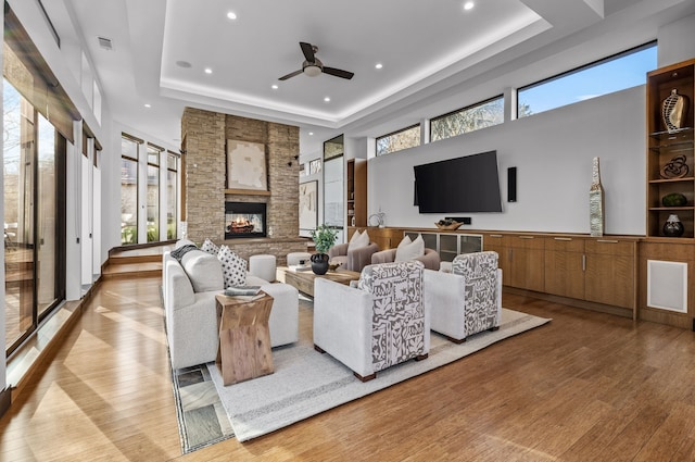 living room featuring a stone fireplace, ceiling fan, a raised ceiling, light hardwood / wood-style flooring, and a high ceiling