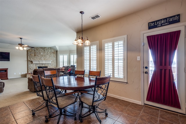 dining area with a stone fireplace, ceiling fan with notable chandelier, plenty of natural light, and dark colored carpet