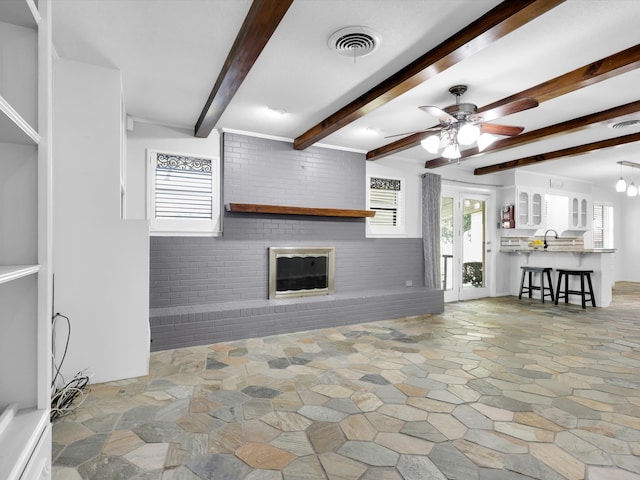 unfurnished living room with ceiling fan, beamed ceiling, brick wall, and a brick fireplace