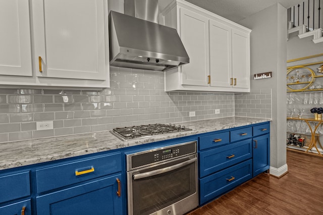 kitchen featuring wall chimney range hood, appliances with stainless steel finishes, white cabinetry, blue cabinetry, and dark wood-type flooring