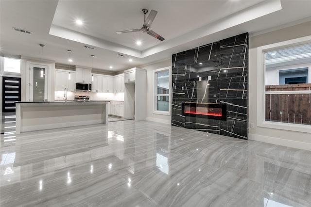 kitchen featuring ceiling fan, a raised ceiling, white cabinetry, a tile fireplace, and pendant lighting