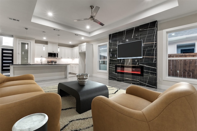 living room with ceiling fan, light wood-type flooring, a fireplace, and a raised ceiling