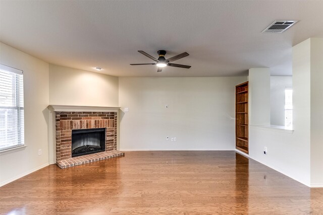 unfurnished living room featuring wood finished floors, baseboards, visible vents, ceiling fan, and a brick fireplace