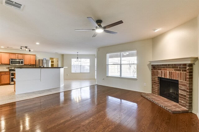 unfurnished living room featuring visible vents, baseboards, light wood-style floors, a brick fireplace, and ceiling fan with notable chandelier