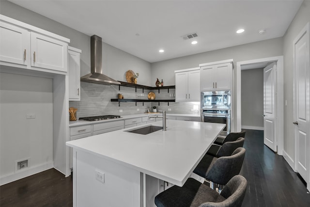 kitchen featuring wall chimney range hood, white cabinets, an island with sink, sink, and stainless steel appliances