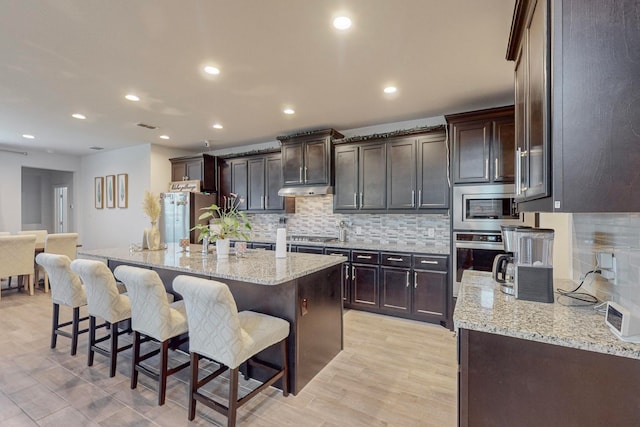 kitchen with stainless steel appliances, light stone counters, light hardwood / wood-style flooring, backsplash, and a center island with sink