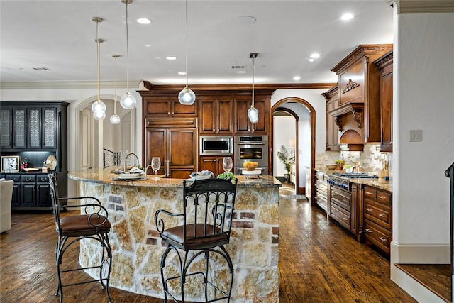 kitchen featuring a kitchen island with sink, built in appliances, hanging light fixtures, light stone counters, and dark hardwood / wood-style flooring