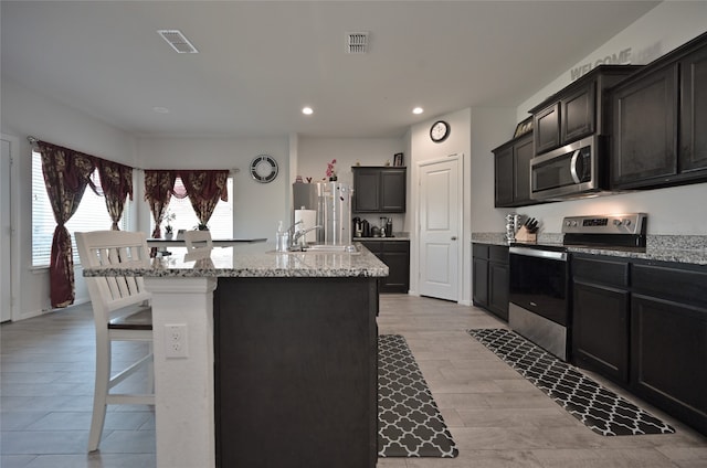 kitchen featuring a kitchen breakfast bar, an island with sink, sink, light stone countertops, and appliances with stainless steel finishes