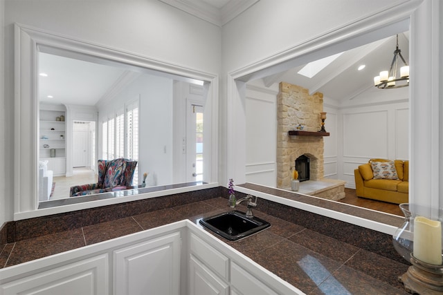 kitchen with sink, a stone fireplace, white cabinetry, vaulted ceiling with skylight, and ornamental molding