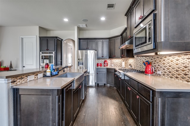 kitchen featuring decorative backsplash, appliances with stainless steel finishes, dark hardwood / wood-style flooring, ventilation hood, and an island with sink