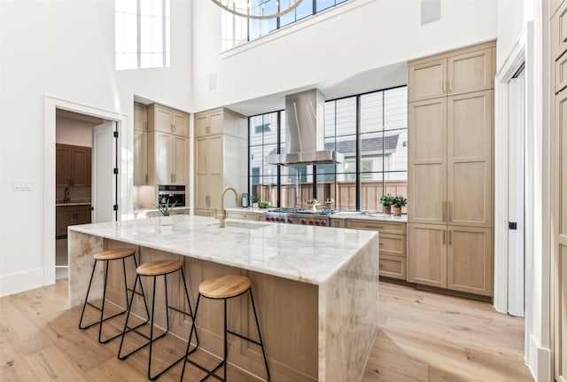 kitchen with sink, a center island with sink, island exhaust hood, light stone countertops, and light hardwood / wood-style floors