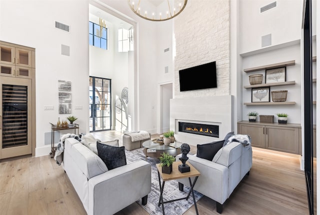 living room featuring wine cooler, a stone fireplace, a notable chandelier, a towering ceiling, and light hardwood / wood-style floors