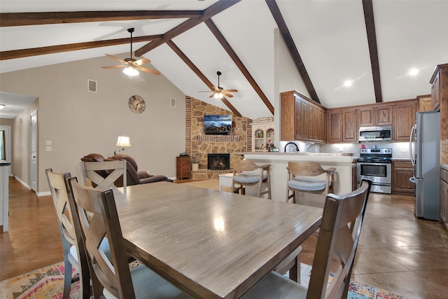 dining space featuring beam ceiling, ceiling fan, high vaulted ceiling, dark tile patterned flooring, and a stone fireplace