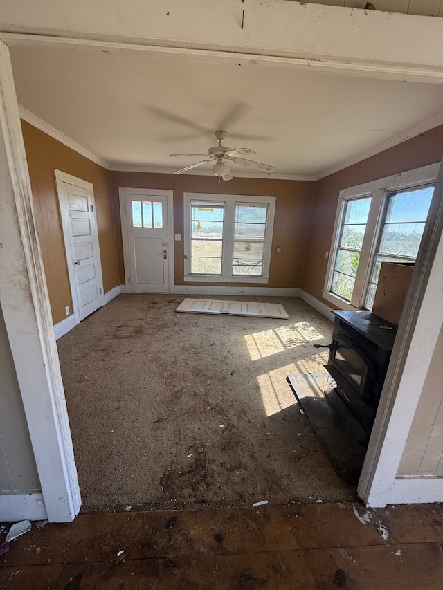 unfurnished living room featuring ceiling fan, a wood stove, ornamental molding, and plenty of natural light