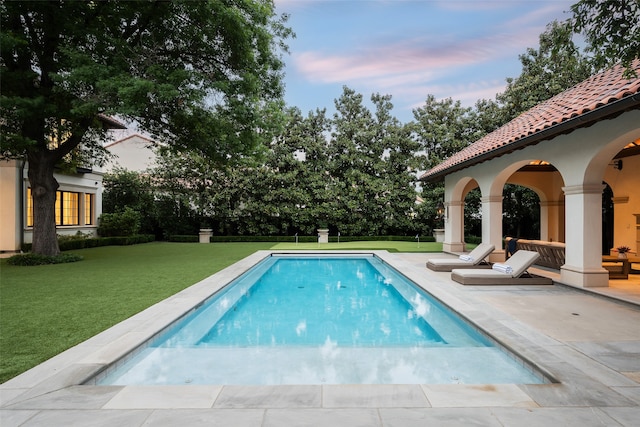 pool at dusk featuring a patio and a lawn