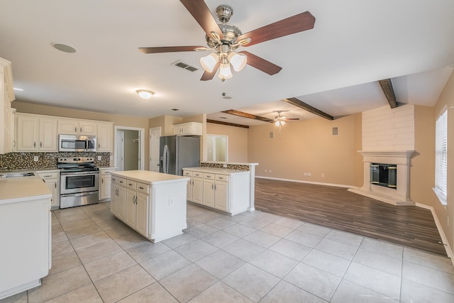 kitchen with beam ceiling, a center island, appliances with stainless steel finishes, a fireplace, and light hardwood / wood-style floors