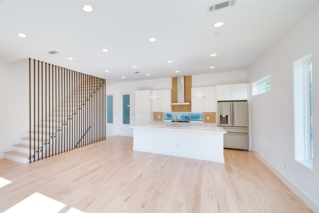 kitchen featuring wall chimney exhaust hood, an island with sink, stainless steel fridge, decorative light fixtures, and white cabinetry