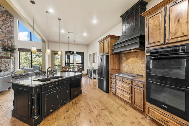 kitchen featuring custom exhaust hood, hanging light fixtures, dark stone counters, black appliances, and light hardwood / wood-style floors