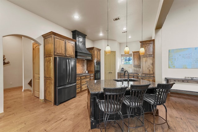 kitchen with light wood-type flooring, stainless steel fridge, pendant lighting, dark stone countertops, and a kitchen island with sink