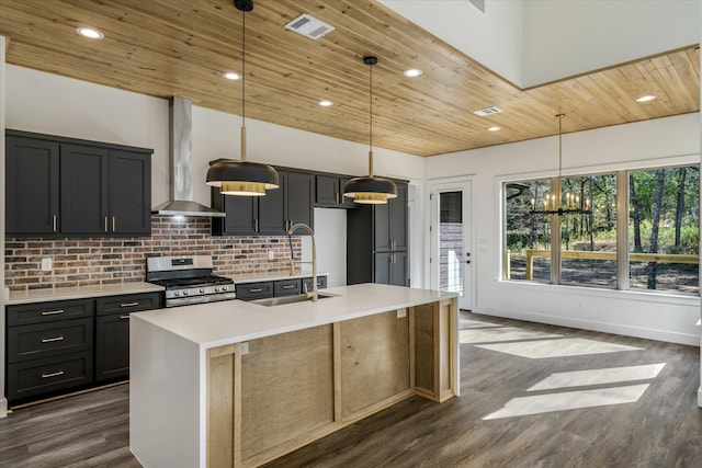 kitchen with gas range, hanging light fixtures, wall chimney range hood, and an island with sink