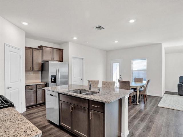 kitchen featuring sink, appliances with stainless steel finishes, dark brown cabinets, light stone countertops, and a center island with sink