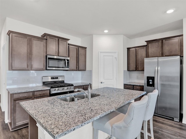 kitchen with light stone counters, a center island with sink, dark hardwood / wood-style floors, stainless steel appliances, and backsplash