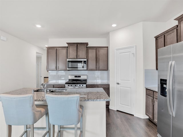 kitchen featuring dark wood-type flooring, appliances with stainless steel finishes, backsplash, light stone countertops, and an island with sink