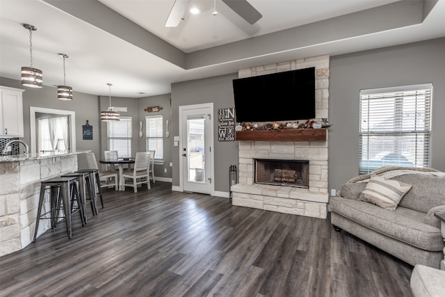 living room with a stone fireplace, ceiling fan, a raised ceiling, and dark hardwood / wood-style flooring