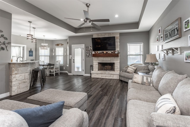 living room with dark wood-type flooring, a fireplace, a tray ceiling, and ceiling fan with notable chandelier