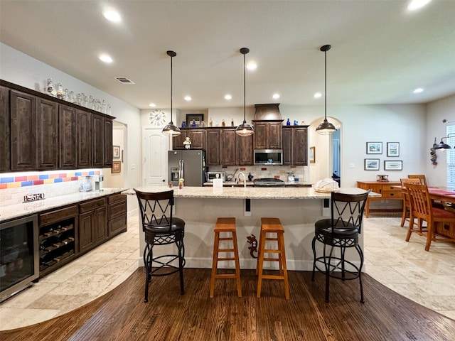 kitchen with beverage cooler, hanging light fixtures, an island with sink, light stone countertops, and stainless steel appliances