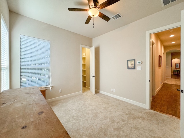 unfurnished bedroom with a walk in closet, multiple windows, light colored carpet, and ceiling fan