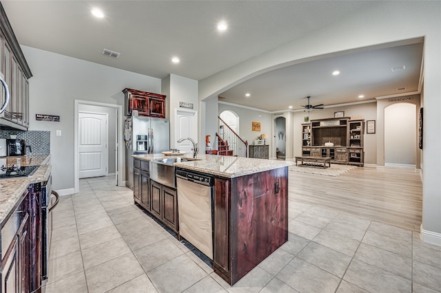 kitchen with light stone counters, stainless steel appliances, ceiling fan, crown molding, and a center island with sink