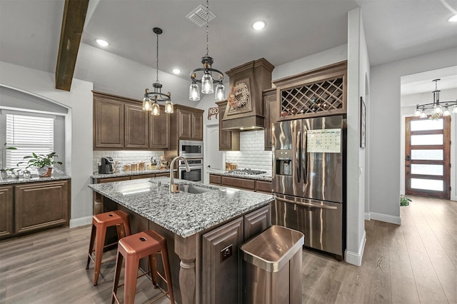 kitchen with light hardwood / wood-style flooring, a center island with sink, appliances with stainless steel finishes, light stone counters, and tasteful backsplash