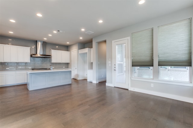 kitchen with wall chimney range hood, dark hardwood / wood-style floors, an island with sink, backsplash, and white cabinetry