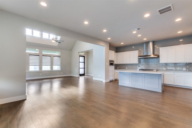 kitchen featuring dark hardwood / wood-style flooring, ceiling fan, wall chimney exhaust hood, white cabinets, and a center island with sink