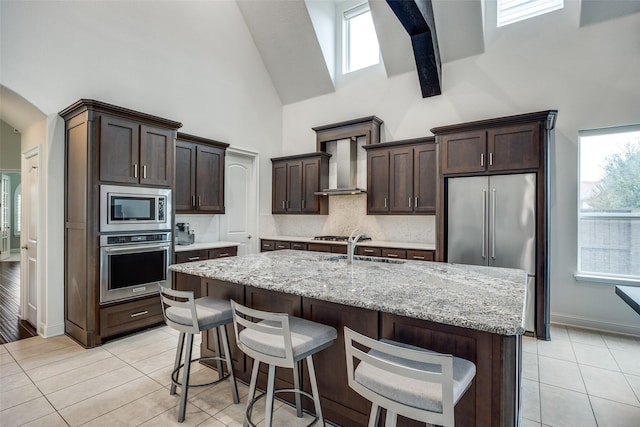 kitchen with sink, a kitchen island with sink, stainless steel appliances, light stone countertops, and wall chimney exhaust hood