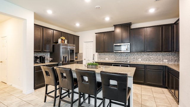 kitchen featuring appliances with stainless steel finishes, a kitchen bar, backsplash, light stone counters, and a kitchen island with sink