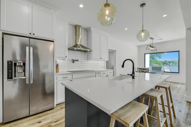 kitchen with white cabinetry, wall chimney range hood, a center island with sink, and stainless steel fridge