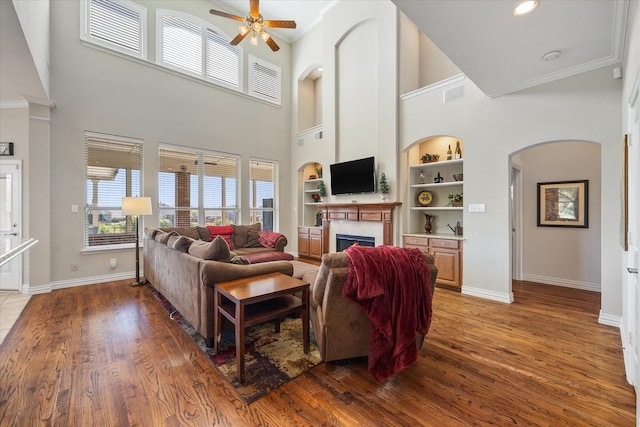 living room featuring a fireplace, built in shelves, ceiling fan, a towering ceiling, and wood finished floors