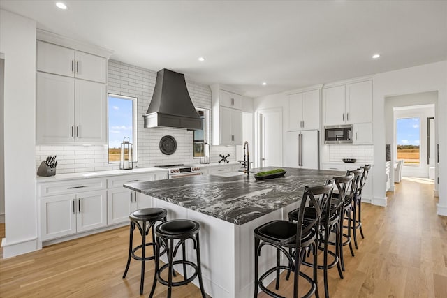 kitchen with built in appliances, light wood-type flooring, wall chimney exhaust hood, an island with sink, and backsplash