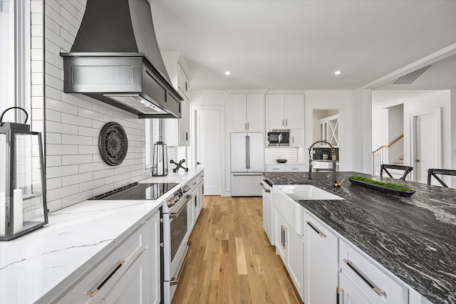 kitchen featuring sink, stone counters, premium range hood, built in appliances, and white cabinets