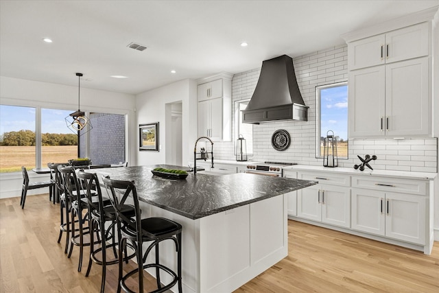 kitchen featuring wall chimney exhaust hood, light wood finished floors, decorative backsplash, and a center island with sink