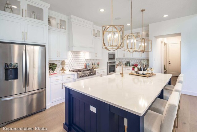 kitchen with appliances with stainless steel finishes, a kitchen island with sink, pendant lighting, light hardwood / wood-style flooring, and white cabinetry