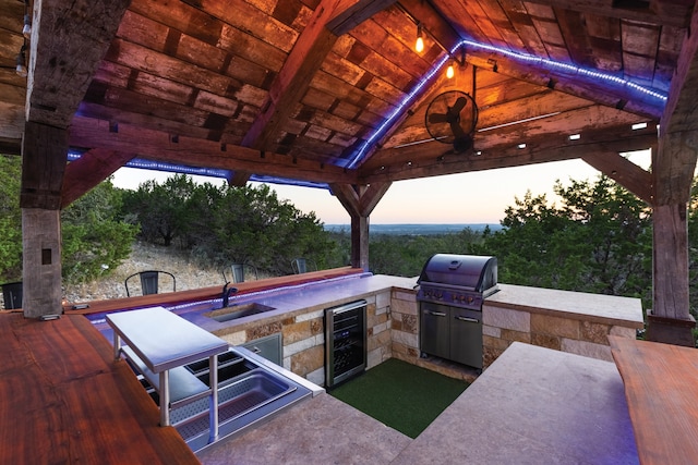 patio terrace at dusk with a grill, area for grilling, wine cooler, sink, and a gazebo