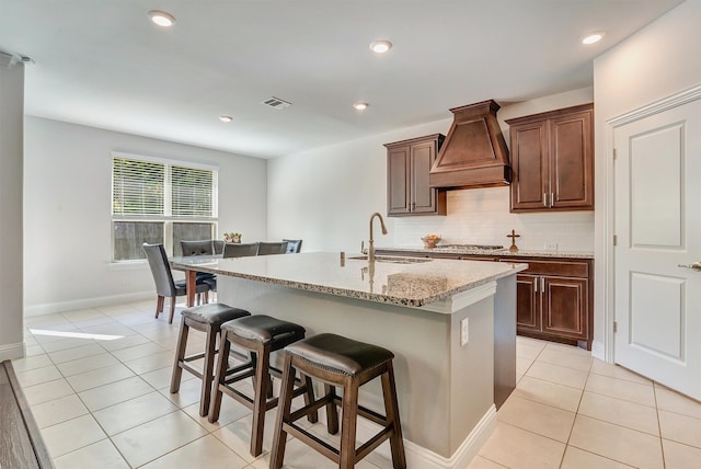 kitchen with light stone countertops, sink, custom exhaust hood, a breakfast bar, and a kitchen island with sink