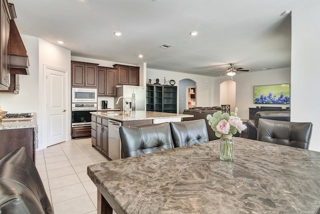 kitchen featuring an island with sink, ceiling fan, stainless steel appliances, dark brown cabinetry, and light stone counters