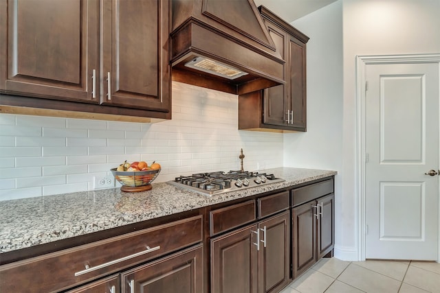 kitchen with stainless steel gas cooktop, light stone countertops, dark brown cabinetry, light tile patterned floors, and custom exhaust hood