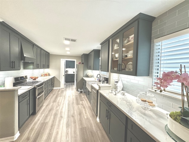 kitchen with tasteful backsplash, wall chimney range hood, light hardwood / wood-style floors, stainless steel appliances, and light stone counters