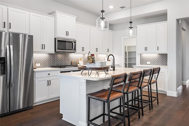 kitchen featuring appliances with stainless steel finishes, hanging light fixtures, white cabinets, a center island with sink, and dark hardwood / wood-style flooring