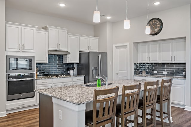 kitchen featuring white cabinets, hanging light fixtures, appliances with stainless steel finishes, light stone countertops, and a kitchen island with sink